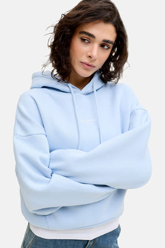 Pull à capuche bleu de JJXX, avec les bras croisés et un jean blanc.