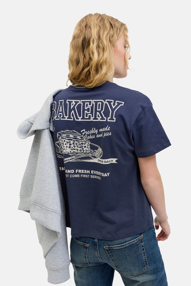 T-shirt bleu à manches courtes de JJXX, avec une impression "Bakery" blanche sur le dos, associé à des jeans et un pull gris.