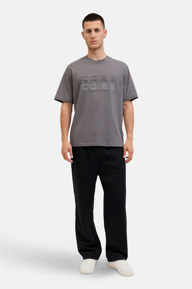 T-shirt gris à manches courtes de CORE by JACK & JONES, avec une impression graphique sur la poitrine, associé à un pantalon noir et des chaussures blanches.