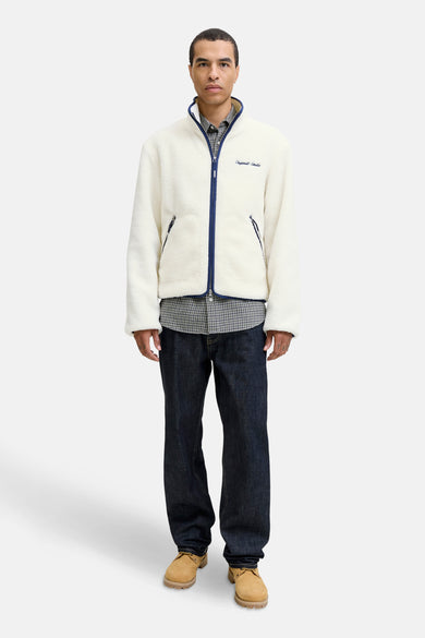 Manteau beige d'ORIGINALS BY JACK & JONES, avec une fermeture éclair bleue et un chemisier à carreaux en dessous, associé à des jeans foncés et des chaussures marron.