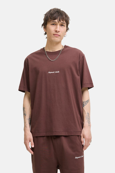 T-shirt marron à manches courtes d'ORIGINALS BY JACK & JONES, avec une impression de texte blanche, porté avec une chaîne argentée.