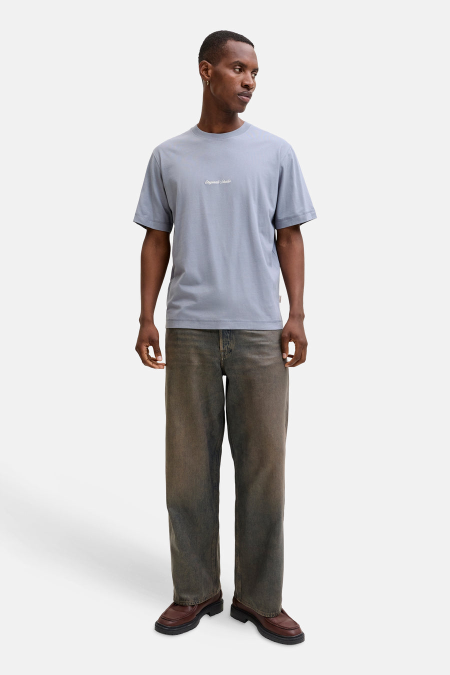 Man in blauwgrijs T-shirt, verkleurde jeans en bruine loafers.