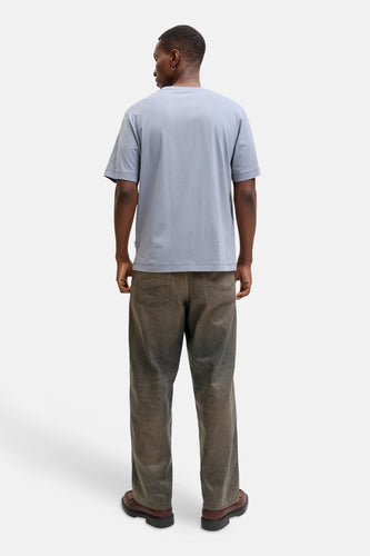 Achterkant van een donkere man in een blauw T-shirt, vuil ogende jeans en bruine schoenen.