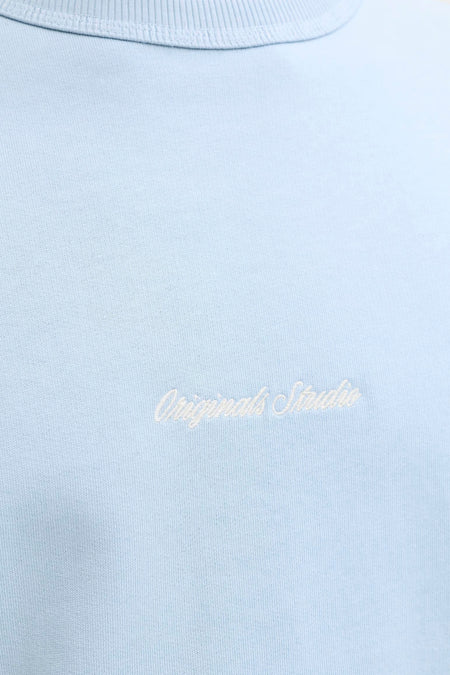 Pull bleu clair à col rond de ORIGINALS BY JACK & JONES avec inscription brodée blanche 'Originals Studio' sur la poitrine.