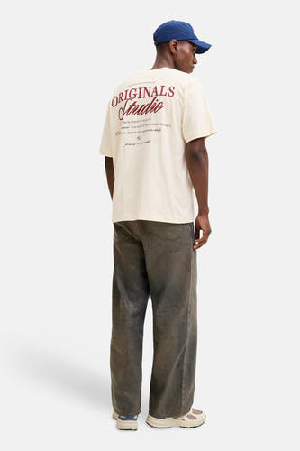 T-shirt écru à manches courtes de ORIGINALS BY JACK & JONES, avec imprimé au dos, porté avec une casquette bleue et un jean gris.