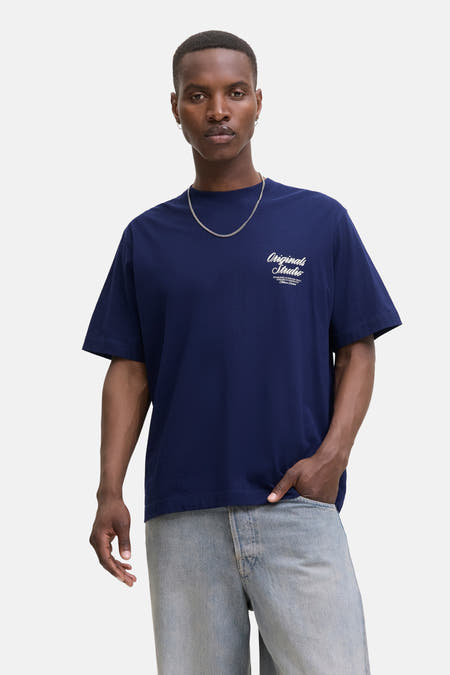 T-shirt bleu à manches courtes d'ORIGINALS BY JACK & JONES, avec du texte blanc sur la poitrine, associé à une chaîne argentée et des jeans.