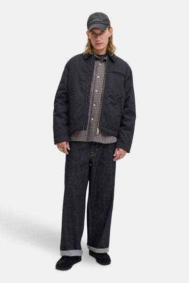 Manteau court noir d'ORIGINALS BY JACK & JONES, porté sur une chemise à carreaux et un jeans foncé, avec un chapeau.
