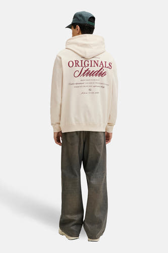 Pull beige à capuche de ORIGINALS BY JACK & JONES, avec imprimé au dos et porté avec une casquette et un jean ample.