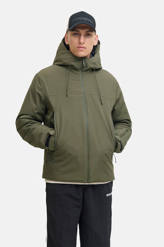 Veste courte verte de CORE BY JACK & JONES avec capuche, portée par un homme avec une casquette noire, un pull de couleur crème, un jogging noir et des baskets beiges.