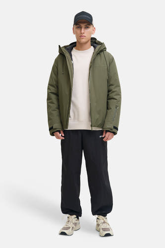 Groene korte jas van CORE BY JACK & JONES met capuchon, gedragen door een man met een zwarte pet, crèmekleurige trui, zwarte joggingbroek en beige sneakers.