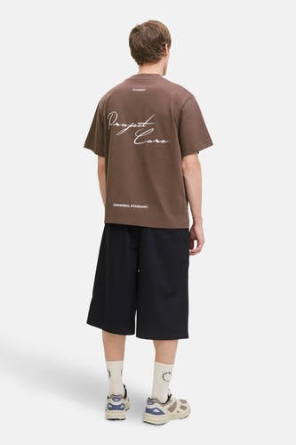 T-shirt brun à manches courtes de CORE BY JACK & JONES, avec une impression de texte blanche sur le dos, porté avec des short noirs.