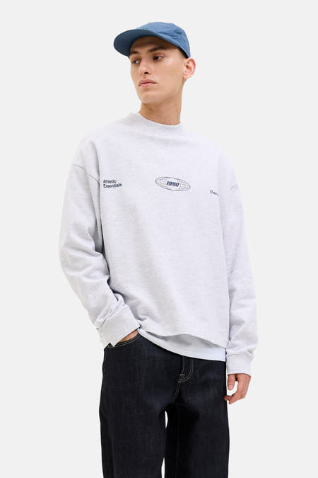 Pull-over gris à col rond de CORE BY JACK & JONES, avec des impressions de logo sur la partie avant, porté avec un chapeau bleu et des jeans foncés.