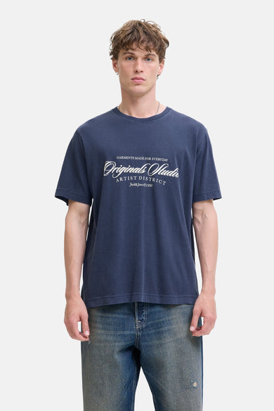 T-shirt bleu à manches courtes d'ORIGINALS par JACK & JONES, avec une impression de texte blanche, porté avec un jean bleu.