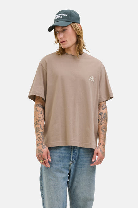T-shirt marron à manches courtes d'Originals by Jack & Jones, avec un petit logo imprimé et porté avec des jeans bleus et une casquette.