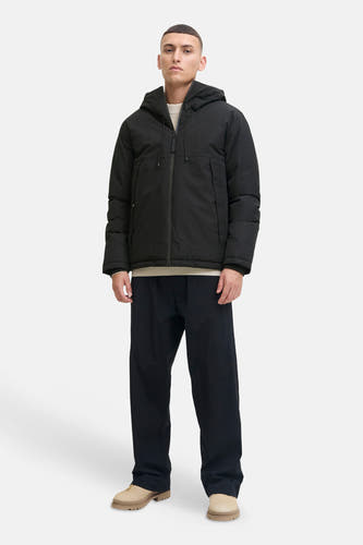 Veste courte noire de CORE BY JACK & JONES avec capuche, fermeture éclair et poches, combinée avec un pull beige clair, un pantalon bleu foncé et des chaussures beiges.