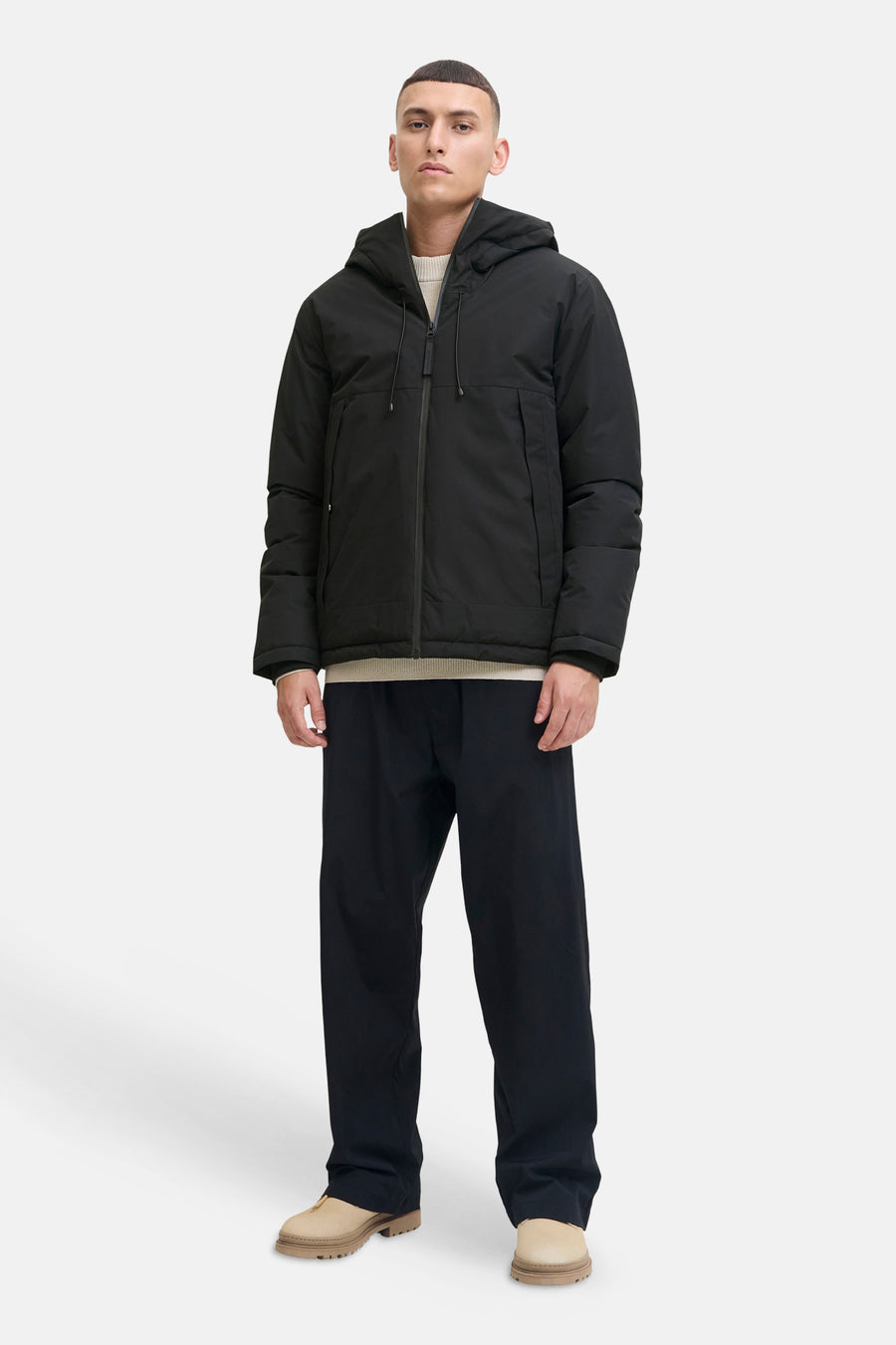 Veste courte noire de CORE BY JACK & JONES avec capuche, fermeture éclair et poches, combinée avec un pull beige clair, un pantalon bleu foncé et des chaussures beiges.