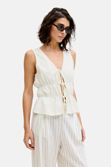 Chemisier blanc sans manches de JJXX avec fermeture à nouer et ourlet péplum, porté avec un pantalon rayé et des lunettes de soleil.