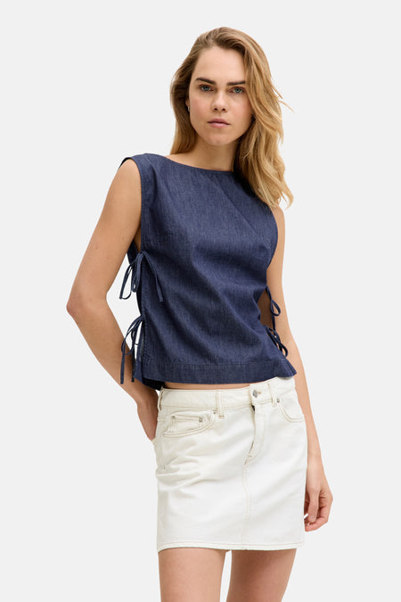 Chemisier sans manches en denim bleu foncé de JJXX avec des nœuds sur les côtés, porté avec une jupe blanche et des mocassins foncés.