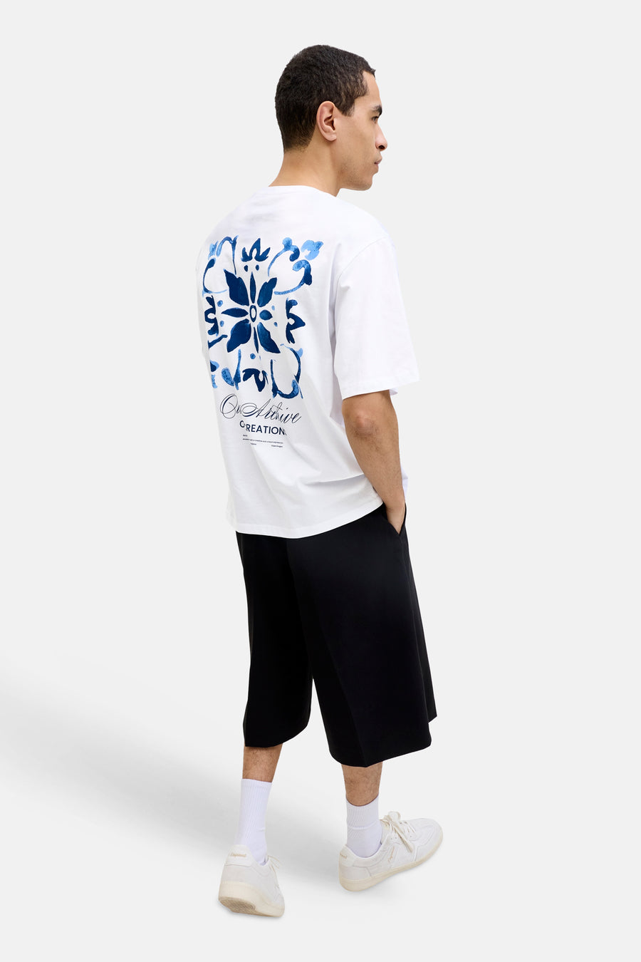 T-shirt blanc ORIGINALS BY JACK & JONES avec motif floral bleu et texte dans le dos, porté avec un short noir et des baskets.