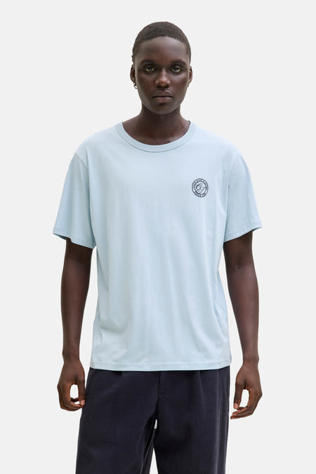 T-shirt bleu à manches courtes d'ORIGINALS BY JACK & JONES, avec logo sur la poitrine, porté avec un pantalon foncé et des chaussures beige.