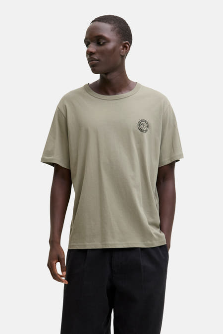 T-shirt vert de mantes courtes man ORIGINALSSSS & J J, petit petit logo sur la poitrine et porté avec un pantalon noir.