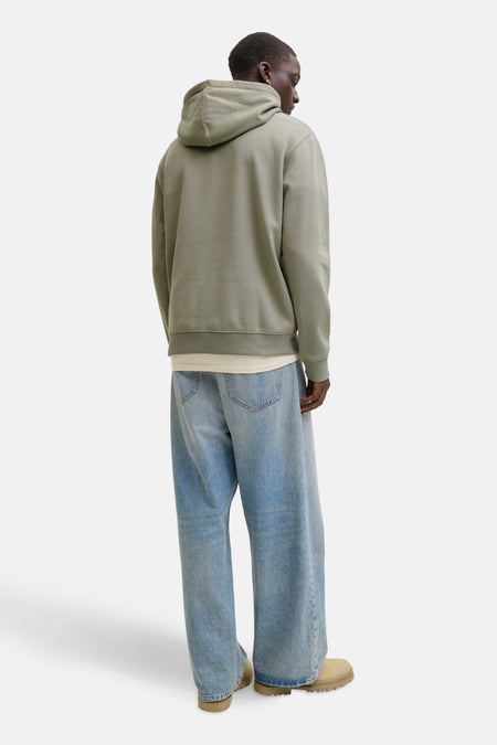 Man van achteren in groene hoodie, wijde jeans en beige schoenen op witte achtergrond.