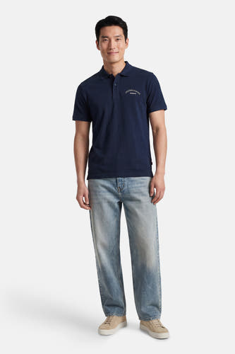 Polo bleu marine de ORIGINALS BY JACK & JONES avec broderie blanche 'JACK&JONES ARCHIVE' sur la poitrine, portée avec un jean clair et des baskets beiges.