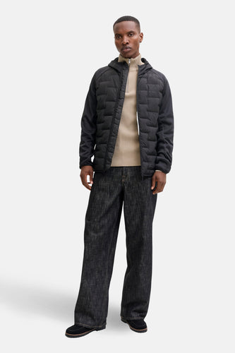 Man in zwarte jas, beige trui en donkere jeans, staand.