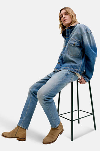 Jeans fuselés bleu moyen en denim de JACK & JONES JEANS INTELLIGENCE, combinés avec une veste en jean et des bottes en daim, assis sur un tabouret.