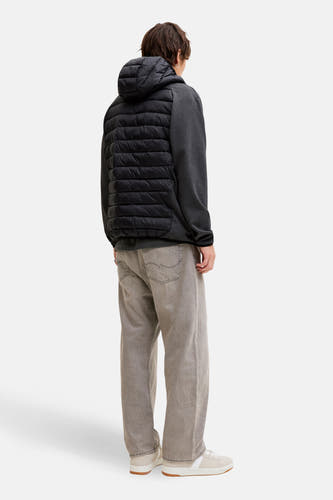 Manteau court noir de CORE BY JACK & JONES, avec une doublure en duvet à l'arrière et un capuchon, porté avec un pantalon gris et des baskets crème.