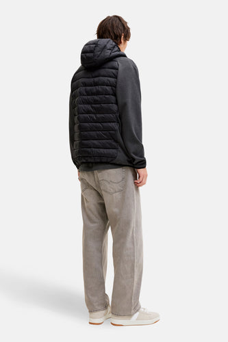 Achteraanzicht van een man in een grijs sweatshirt met een zwart pufferjack met capuchon, grijze jeans en beige sneakers.