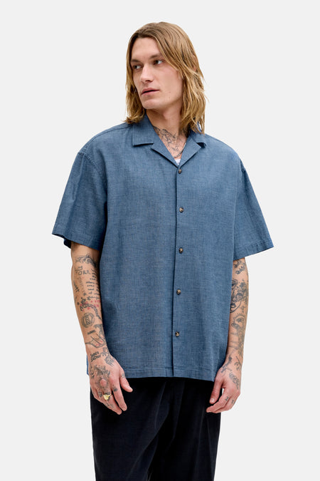 Chemise bleue à manches courtes de PREMIUM by JACK & JONES, avec fermeture boutonnée et portée avec un pantalon noir et des mocassins marron.