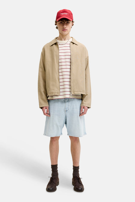 Jeansshort van lichtblauw denim van JACK & JONES JEANS INTELLIGENCE, met gerafelde randen, gedragen met een beige jas en een gestreepte top.
