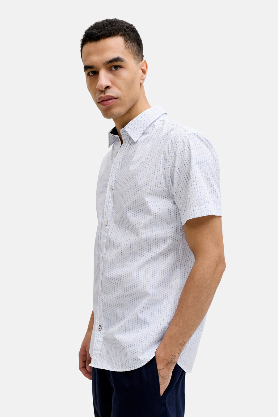 Vue de côté d'une chemise blanche à manches courtes avec motif à pois bleus de PREMIUM by JACK & JONES, portée par un mannequin avec un tatouage sur le bras et la main dans la poche du pantalon.