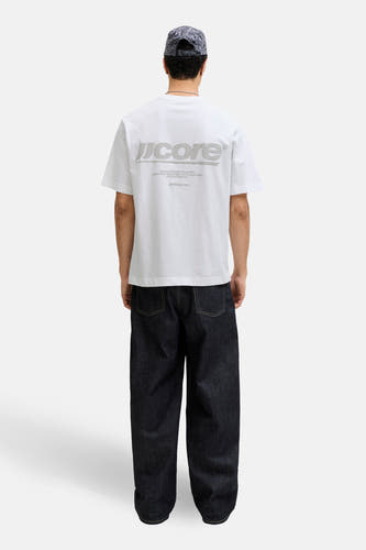 T-shirt blanc à manches courtes de CORE BY JACK & JONES, avec un imprimé logo au dos, combiné avec un jean foncé et une casquette.