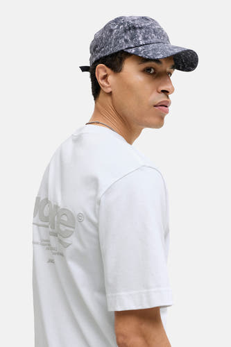 T-shirt blanc à manches courtes de CORE BY JACK & JONES, avec logo de marque sur le dos, porté avec un chapeau.