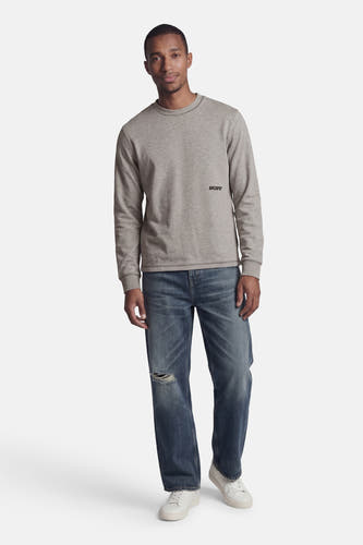 Pull gris à col rond de CORE BY JACK & JONES, avec logo noir 'JJCORE' sur la poitrine, porté par un homme avec un jean déchiré et des baskets blanches.