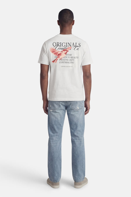T-shirt blanc à manches courtes de ORIGINALS BY JACK & JONES, avec imprimé d'oiseau au dos, combiné avec un jean bleu et des chaussures en daim.
