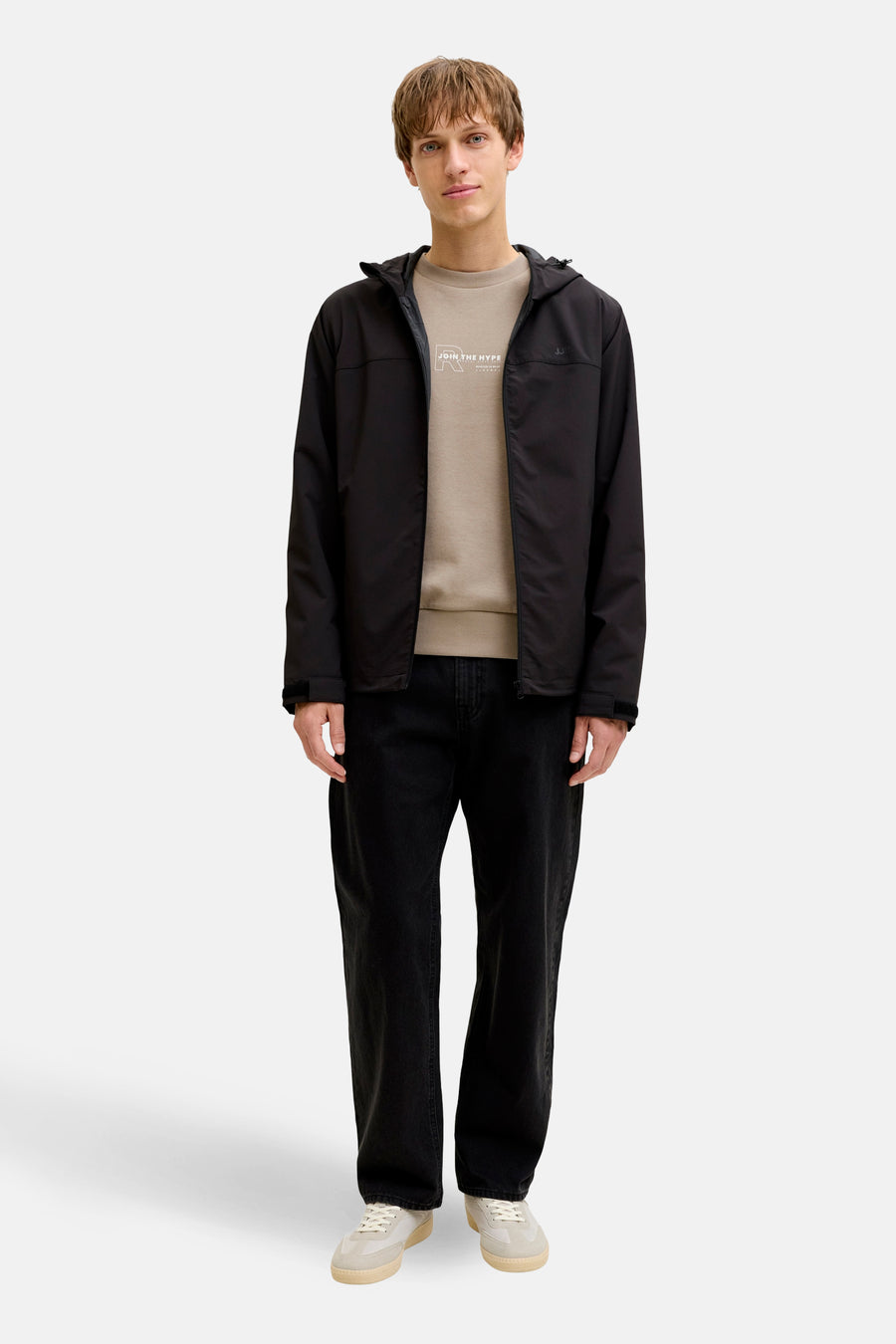 Bruine sweater (ronde hals) van JJREBEL, gedragen onder een zwarte jas met capuchon en donkere jeans.