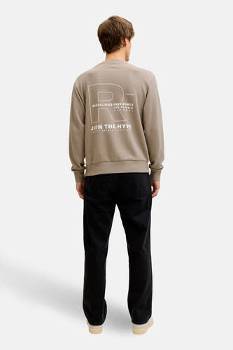 Pull marron à col rond de JJREBEL, avec inscription blanche dans le dos, combiné avec un jean noir et des baskets de couleur crème.
