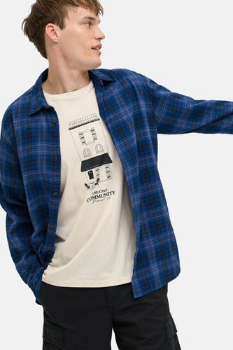 T-shirt beige à manches courtes JJREBEL, avec un imprimé graphique d'un bâtiment, porté sous une chemise à carreaux bleus.