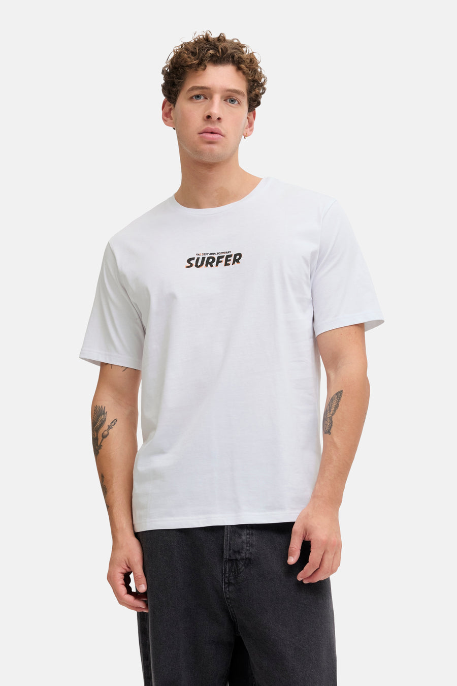 T-shirt blanc à manches courtes de JJREBEL, avec un imprimé texte sur la poitrine, combiné avec un jean foncé et des baskets claires.