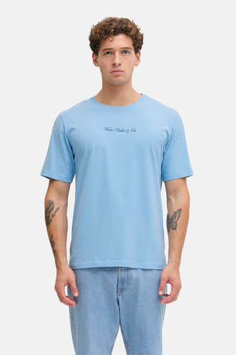 T-shirt bleu à manches courtes de JJREBEL, porté sous une chemise bleu marine avec un jean clair et des chaussures écru.