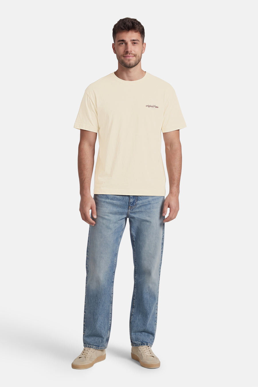 T-shirt beige de ORIGINALS BY JACK & JONES avec imprimé 'originals © 1990', porté par un homme avec un jean à boutons latéraux et des baskets beiges perforées.