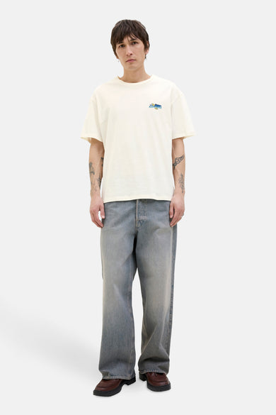 T-shirt blanc à manches courtes d'ORIGINALS BY JACK & JONES, avec petit logo, porté avec des jeans décontractés et des chaussures marrons.