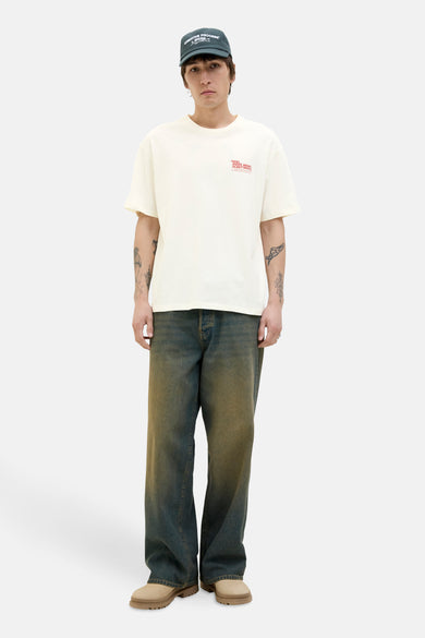T-shirt blanc à manches courtes d'ORIGINALS BY JACK & JONES, avec un petit imprimé rouge et porté avec des jeans décontractés et des chaussures beige.