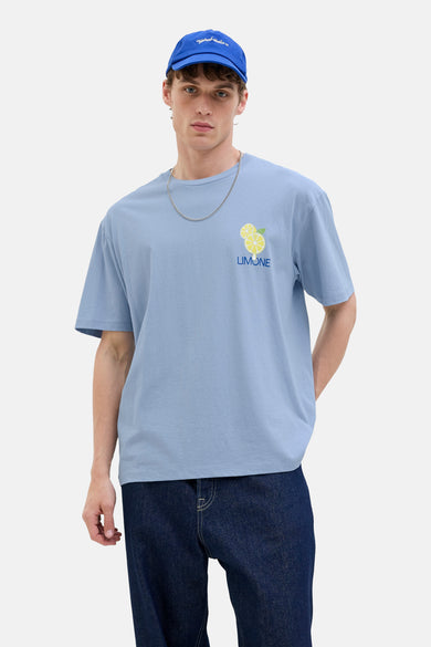 T-shirt bleu à manches courtes d'Originals by Jack & Jones, avec imprimé citron, porté avec des jeans bleus marine et un chapeau.