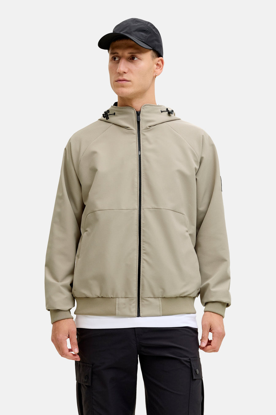 Manteau court beige de CORE BY JACK & JONES, avec fermeture éclair et capuche, combiné avec un pantalon noir et une casquette.