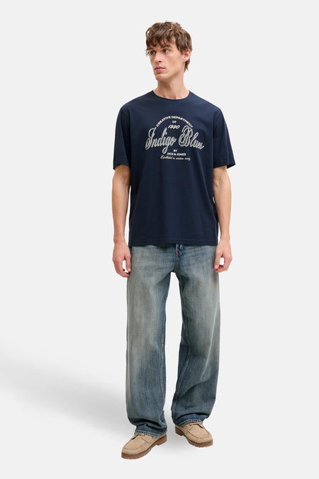 Man met marineblauw T-shirt, wijde jeans en bruine loafers op een witte achtergrond.