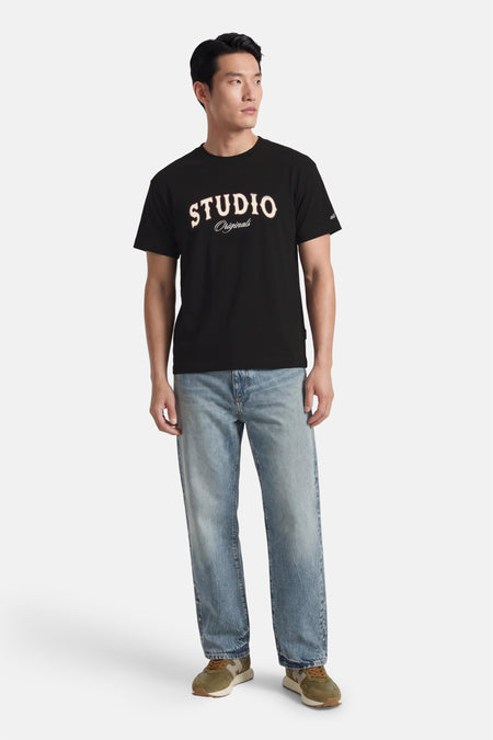T-shirt noir à manches courtes de ORIGINALS BY JACK & JONES, avec imprimé texte blanc "STUDIO" et combiné avec un jean et des baskets vertes.